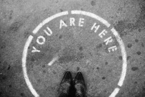 Person wearing black leather shoes standing on a painted circle with "You Are Here" written on the asphalt ground